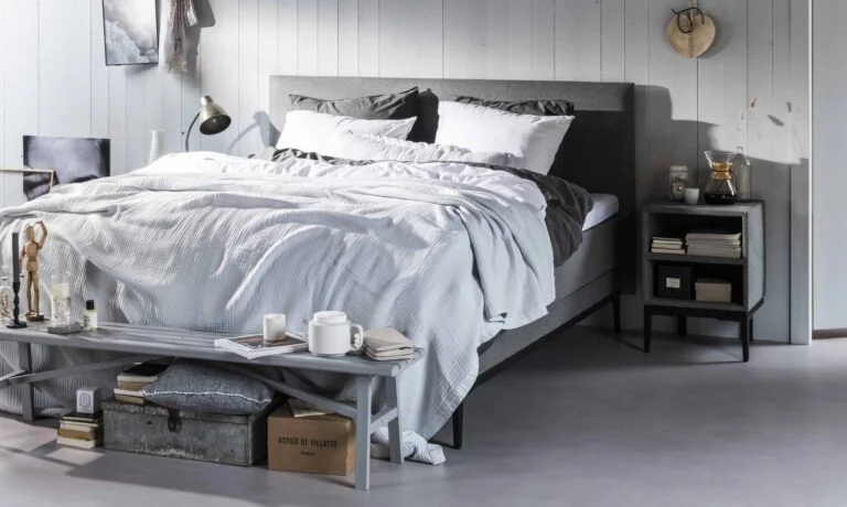 Een moderne slaapkamer met een stijlvol grijs boxspringbed, opgemaakt met een wit en grijs dekbedovertrek en meerdere kussens. Voor het bed staat een klein houten tafeltje met een kopje thee, een boek en een opgevouwen deken. De ruimte heeft een rustige en minimalistische uitstraling met een lichte houten wand en een industriële vloerlamp op de achtergrond.