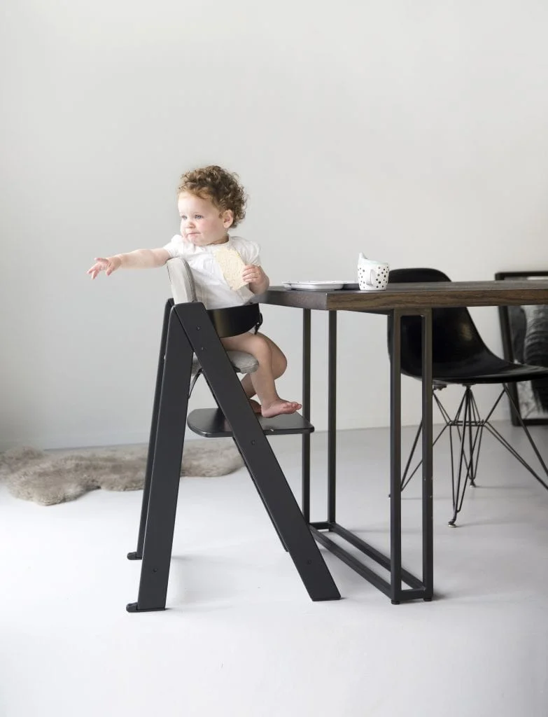 Een peuter met krullend haar zit in een moderne, zwarte houten kinderstoel aan een donkere eettafel. Het kind draagt een wit shirt en heeft een koekje in de hand terwijl het met de andere hand iets probeert te pakken. De ruimte heeft een minimalistisch interieur met een witte vloer en muur, en zwarte designstoelen bij de tafel.