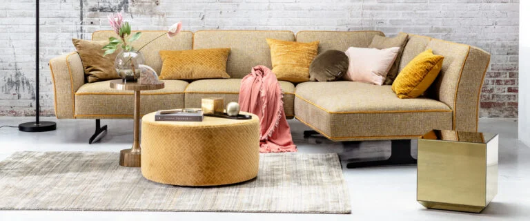 Een sfeervolle woonkamer met een grote hoekbank in een warme gele tint, aangevuld met bijpassende kussens. Voor de bank staat een ronde poef met een luxe textuur, gebruikt als salontafel met decoratieve accessoires. Een roze plaid ligt speels over de bank, terwijl een zacht vloerkleed de ruimte een gezellige uitstraling geeft.