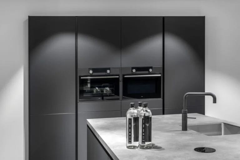 Een moderne keuken met strakke, matzwarte kasten en ingebouwde ovens. Het keukenblad heeft een betonlook afwerking en is voorzien van een minimalistische zwarte kraan. Op het aanrecht staan twee stijlvolle waterflessen, wat bijdraagt aan de luxe en industriële uitstraling van de keuken.