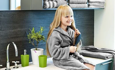 Een jong meisje met lang blond haar zit in een badkamer en borstelt glimlachend haar haar. Ze draagt een zachte, grijze badjas. Op de achtergrond is een blauwe tegelmuur te zien, samen met een witte wastafel en een groene plant in een witte pot. Een groene beker staat op het aanrecht.