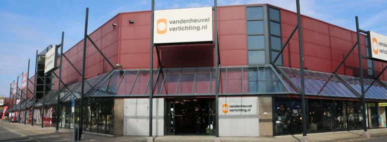 Vooraanzicht van een Van den Heuvel Verlichting-winkel met een roodbruine gevel en grote glazen entree. Boven de ingang hangt een wit bord met het logo en de website 'vandenheuvelverlichting.nl'. Het interieur van de winkel is gedeeltelijk zichtbaar door de open deuren.