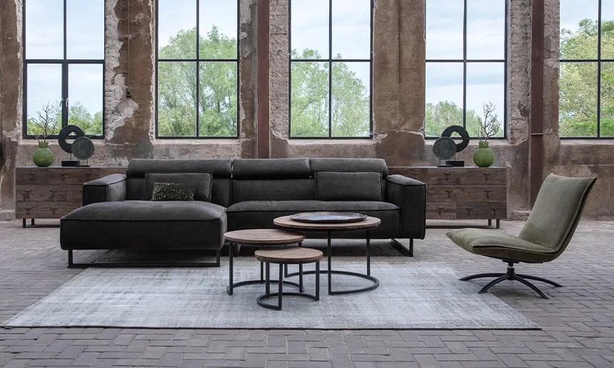 Een industriële woonkamer met een donkergrijze hoekbank en een set ronde salontafels met houten bladen en metalen onderstellen. De ruimte heeft een robuuste uitstraling met grote ramen, verweerde bakstenen muren en een betonnen vloer. Een licht vloerkleed ligt onder de meubels, wat zorgt voor contrast en warmte in het interieur.