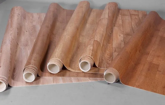 Drie rollen vinyl vloerbedekking met een houtlook, gedeeltelijk uitgerold op een betonnen ondergrond. De vloer heeft een warme, natuurlijke houtstructuur en biedt een praktische en stijlvolle oplossing voor diverse interieurstijlen.