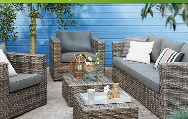 Een sfeervolle tuinset van grijs wicker met donkergrijze kussens, bestaande uit een loungesofa, een fauteuil en een salontafel. De salontafel is gedecoreerd met een dienblad, kaarsen en een vaas met bloemen. Op de achtergrond is een blauwe houten wand te zien, wat zorgt voor een frisse en moderne uitstraling.