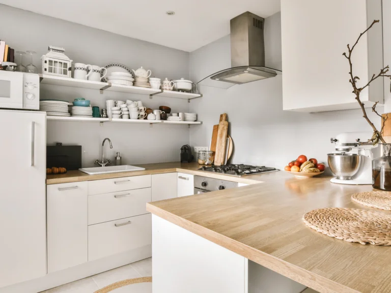 Een lichte, moderne keuken met witte kasten en een houten werkblad. Boven het aanrecht hangen open planken met netjes gestapeld servies en keukengerei. De keuken is voorzien van een gaskookplaat met een rvs-afzuigkap, een spoelbak en stijlvolle houten snijplanken tegen de muur. Verse tomaten en een gevlochten onderzetter zorgen voor een warme, huiselijke sfeer.