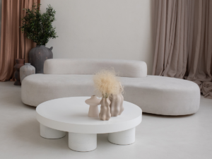Een minimalistische woonkamer met een organisch gevormde beige bank en een ronde witte salontafel. Versierd met beige vazen, pluizig drooggras en zachte gordijnen op de achtergrond. Ein minimalistisches Wohnzimmer mit einem organisch geformten beigen Sofa und einem runden weißen Couchtisch. Dekoriert mit beigen Vasen, flauschigem Trockengras und weichen Vorhängen im Hintergrund.
