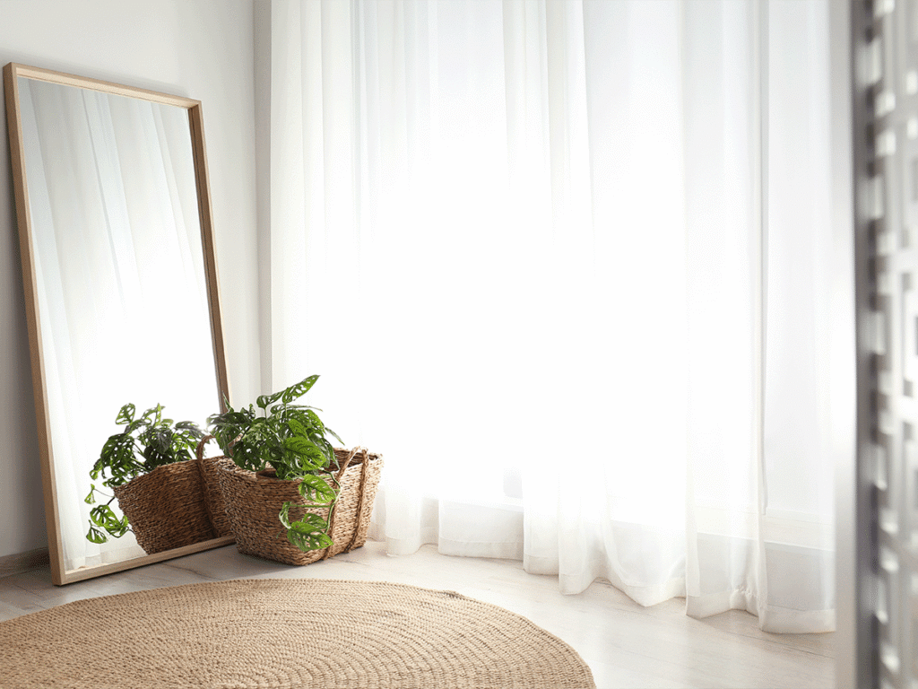 Licht interieur met een grote houten passpiegel, een ronde jute mat en een kamerplant in een mand voor een raam met witte vitrage.