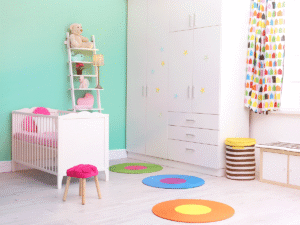Een moderne, kleurrijke babykamer met een witte houten wieg en een grote witte kastenwand versierd met sterren. De kamer heeft een mintgroene accentmuur, een witte decoratieve ladder en drie ronde, felgekleurde vloerkleden op een lichte vloer.