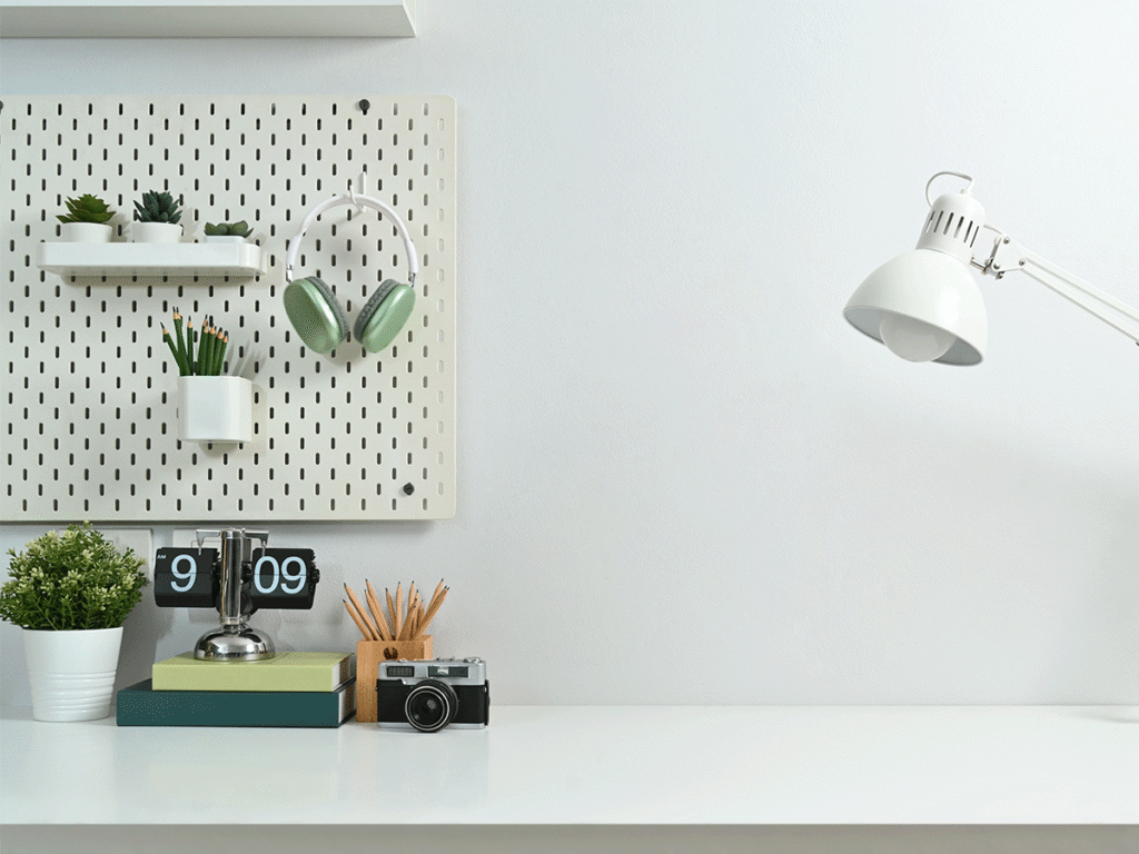 Strak ingericht wit bureau met een gatenbord aan de muur waaraan plantjes en een groene koptelefoon hangen. Op het bureau staan boeken met een retro klok, een potlodenhouder, een vintage camera en een witte bureaulamp.